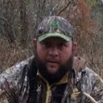 Chris Cook dressed in a camo hat and jacket holding his hunt (deer).